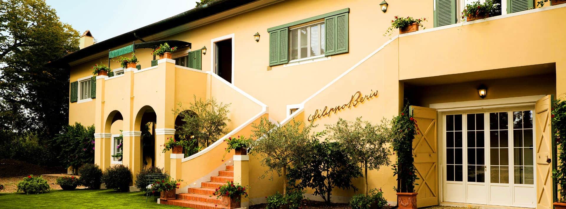 Casa con facciata gialla, finestre verdi e scala esterna, circondata da piante e fiori.